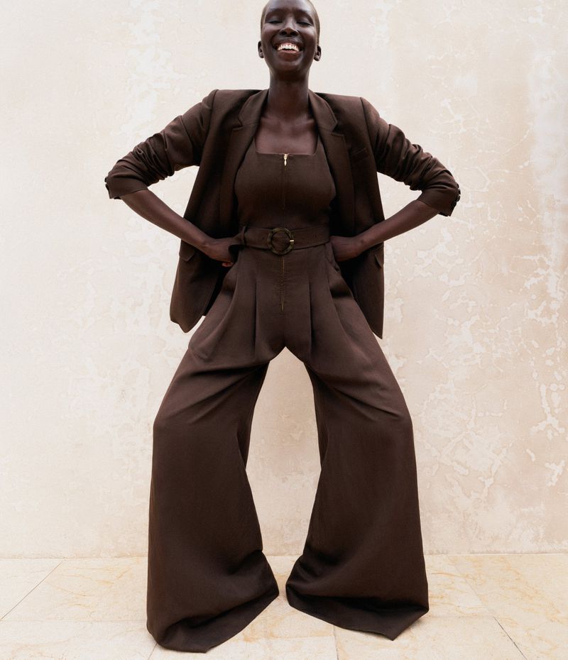 Smiling African woman poses in a studio wearing a brown suit, brown top, and palazzo pants against a neutral backdrop, fashion shot.