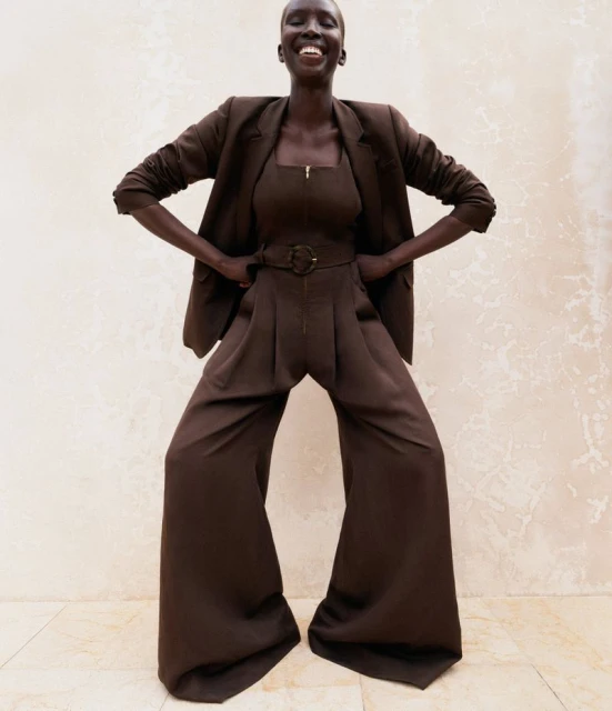Smiling African woman poses in a studio wearing a brown suit, brown top, and palazzo pants against a neutral backdrop, fashion shot.