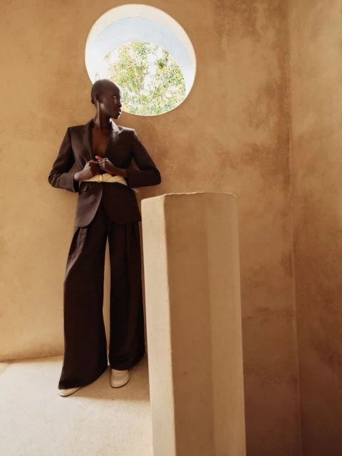 An African woman in a brown suit poses in an earth-toned room by a round window; fashion photography, style, and beauty concept.