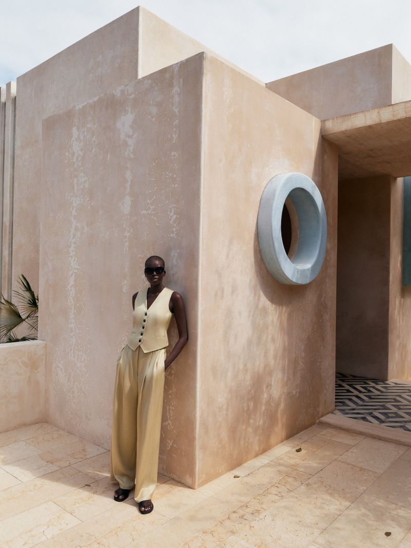Woman in vest and wide-leg pants poses near the corner of a light-beige building, wearing sunglasses, with a patterned floor.