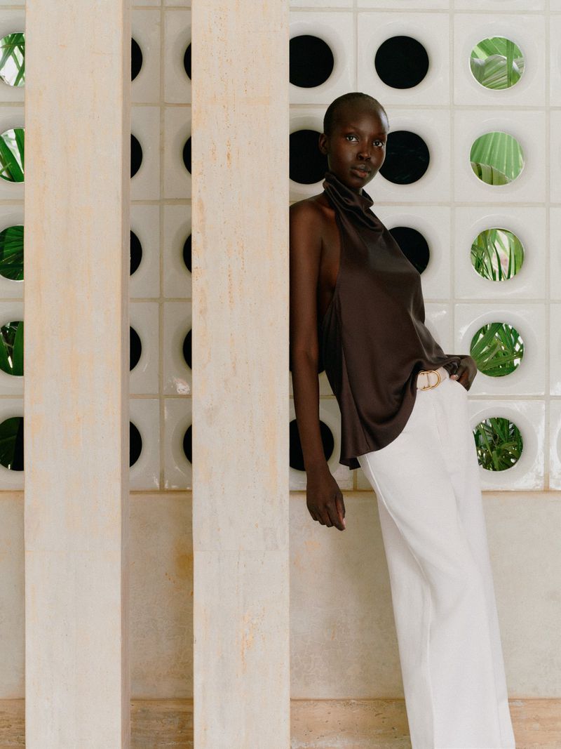 Elegant African-American woman in brown halter top and white wide-leg pants poses indoors near tiled wall and decorative window openings.