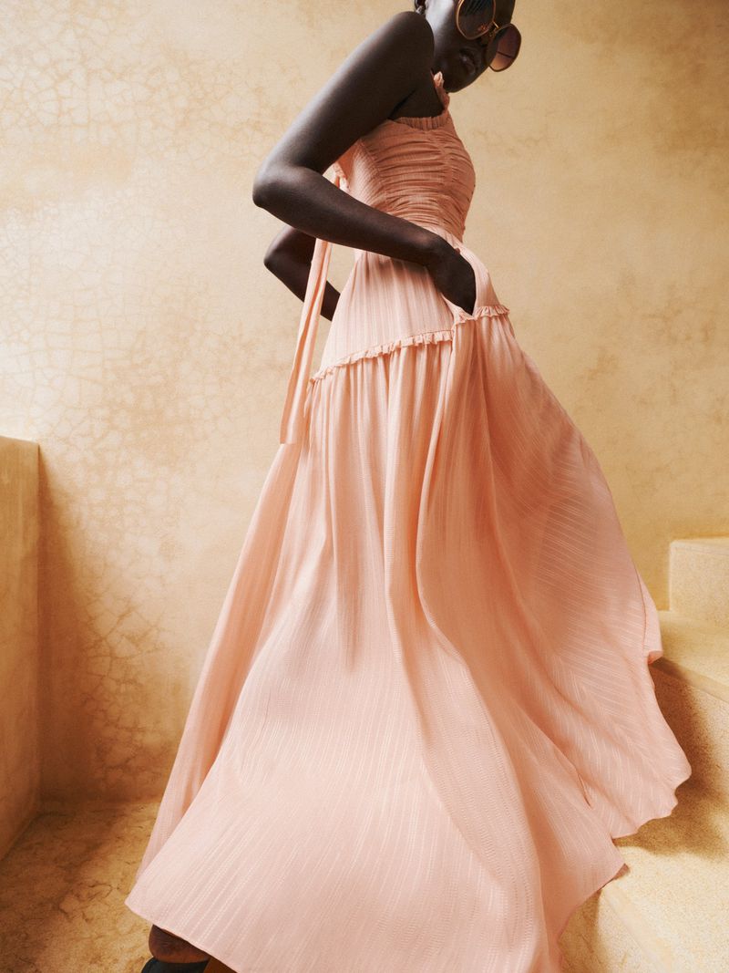An adult woman in a long pink dress with her hands in her pockets stands on stairs in a fashion-editorial style portrait. 