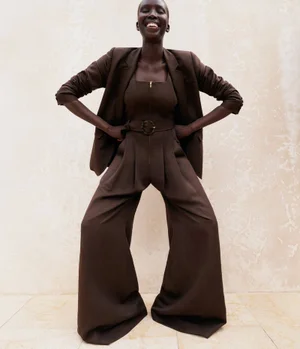 Model Ajok Madel wearing a dark brown blazer and wide-leg trousers against a neutral textured wall.