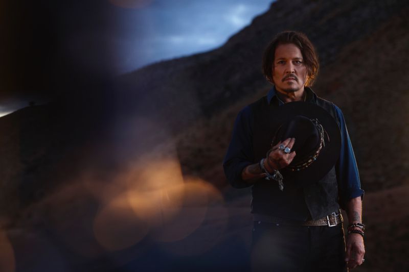Portrait of a light-skinned adult male with medium brown hair, wearing a black vest and jeans, holding a black hat in an outdoor landscape setting.