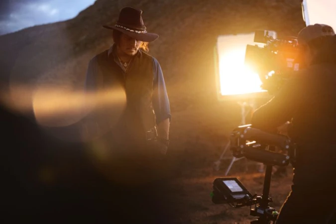 Adult male wearing a cowboy hat and vest stands outdoors. Camera and lighting equipment are visible, suggesting a behind-the-scenes look.