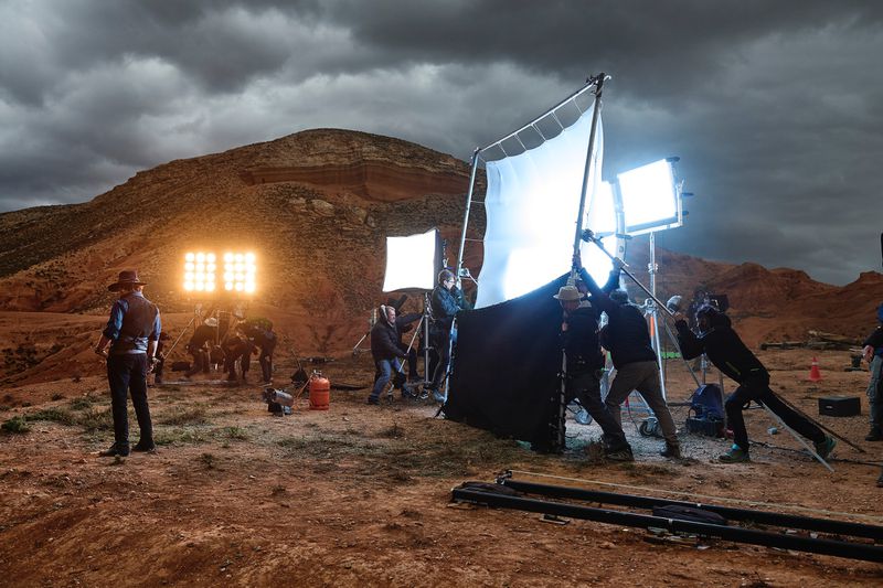 Film set in a desert environment with a lone figure in Western attire, lighting equipment, and a film crew under a cloudy sky.