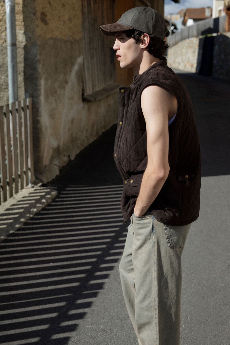 Young man stands on a road in olive-green cap, brown vest, and khaki pants. Sunlight and shadow patterns on the asphalt street.