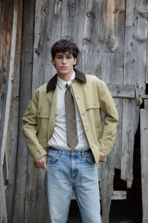 Young man poses in front of rustic barn, wearing a light brown jacket, striped shirt, blue jeans and patterned tie in an outdoor setting.