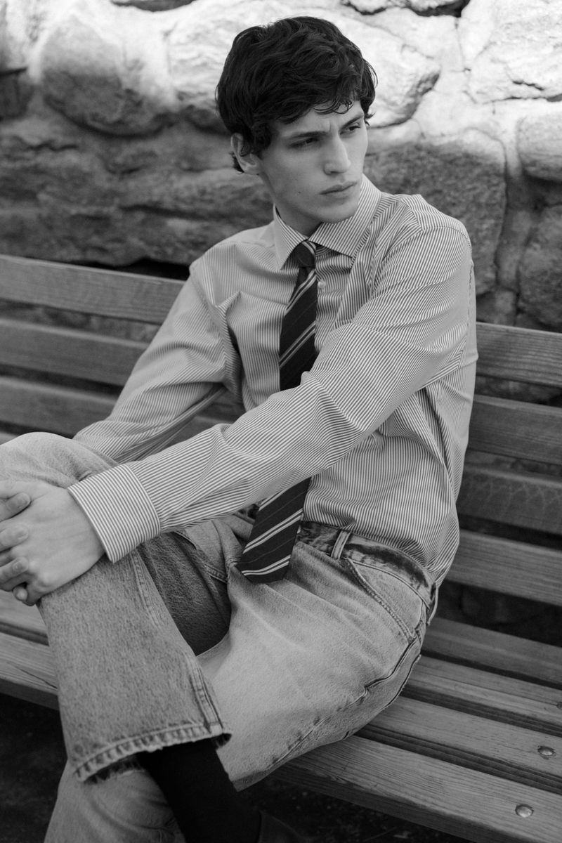 Black and white shot of a young adult male sitting on a park bench wearing jeans, striped shirt, and tie in an outdoor setting.