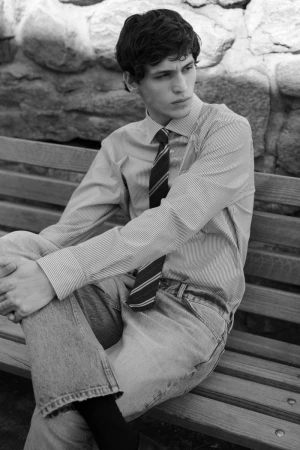 Black and white shot of a young adult male sitting on a park bench wearing jeans, striped shirt, and tie in an outdoor setting.