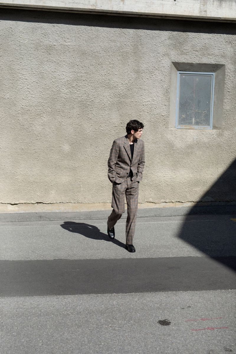 A man in a brown suit walks on an asphalt road with a building in the background on a sunny day, casting strong shadows.