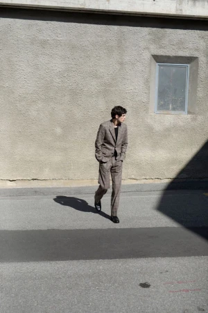 A man in a brown suit walks on an asphalt road with a building in the background on a sunny day, casting strong shadows.