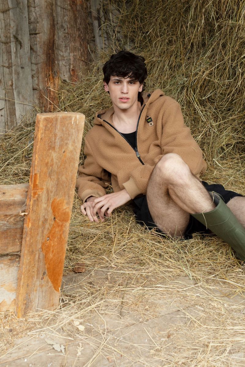 A young adult male rests in a pile of hay wearing a tan hooded jacket, black shorts, and green boots, posing beside a wooden structure.