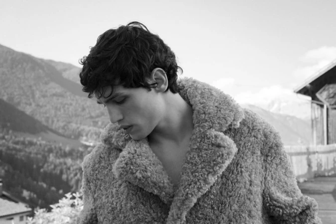 Monochrome portrait of a young adult male wearing a shaggy coat, set against a background of mountains and trees under a bright sky.