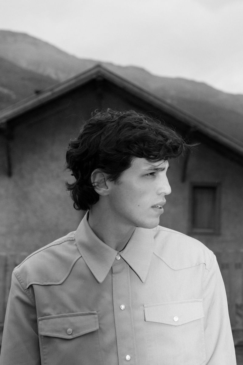 Monochrome portrait of a young adult man in a Western shirt against a backdrop of a rural house and distant mountains.