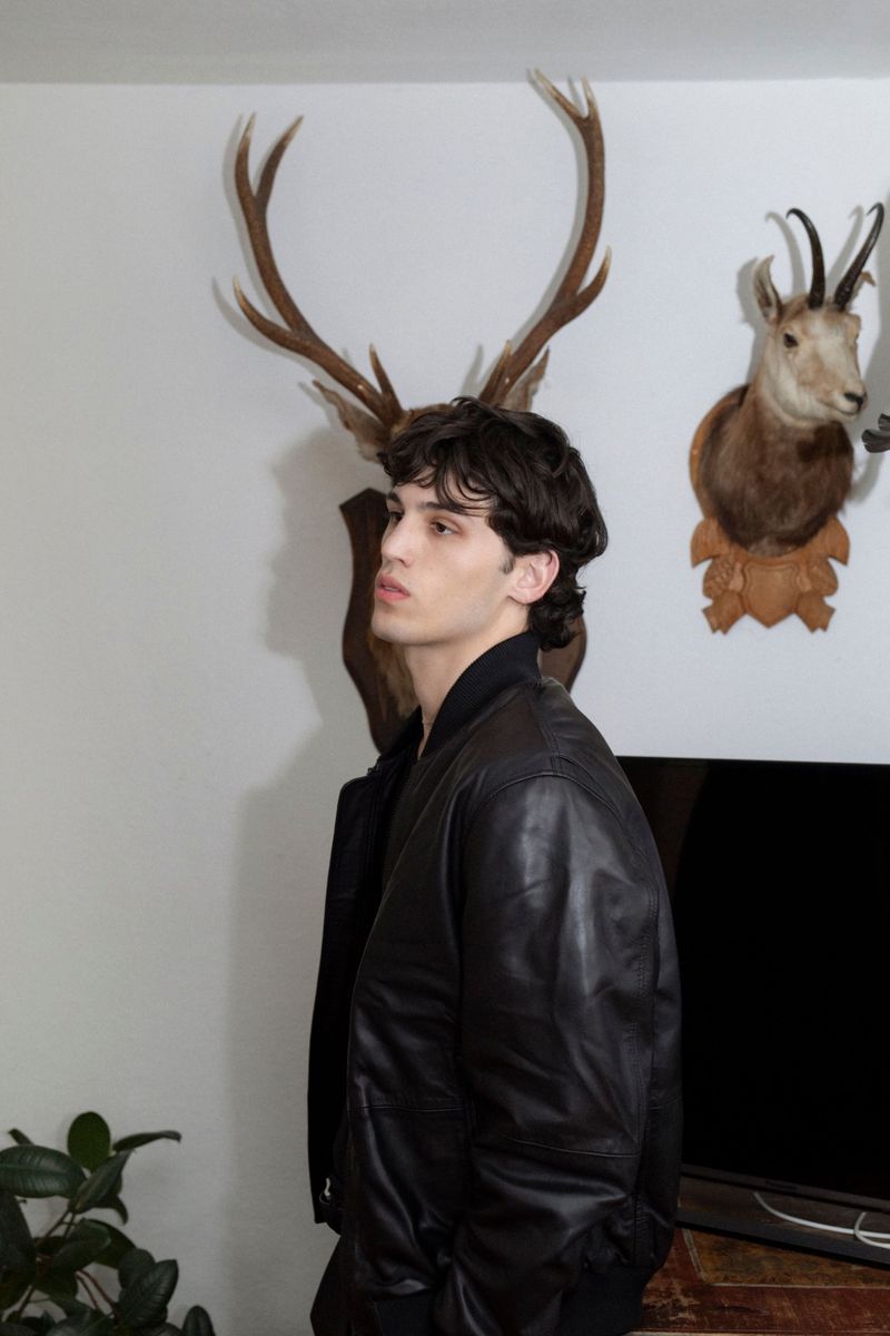 Man in leather jacket poses indoors near mounted animal heads. Fashion shot with dark hair, pale skin and a leather jacket.