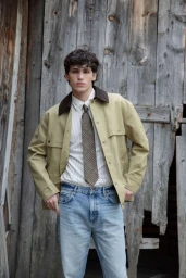 A young male model posing against a rustic weathered wooden wall wearing a khaki jacket and blue jeans.