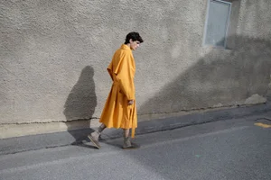 Model wearing a vibrant yellow trench coat walking past a textured concrete wall for GQ Germany