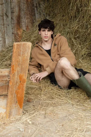 Model Santiago Rivata reclined in straw wearing a camel fleece hoodie and green rubber boots in a rustic barn for GQ Germany.