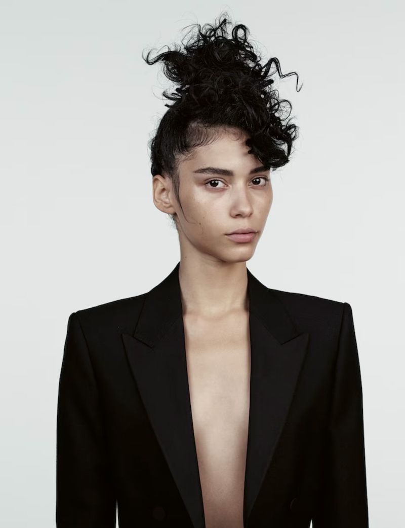 Portrait of a woman in a black blazer, featuring a unique hairstyle and minimalistic makeup, set against a stark white backdrop.