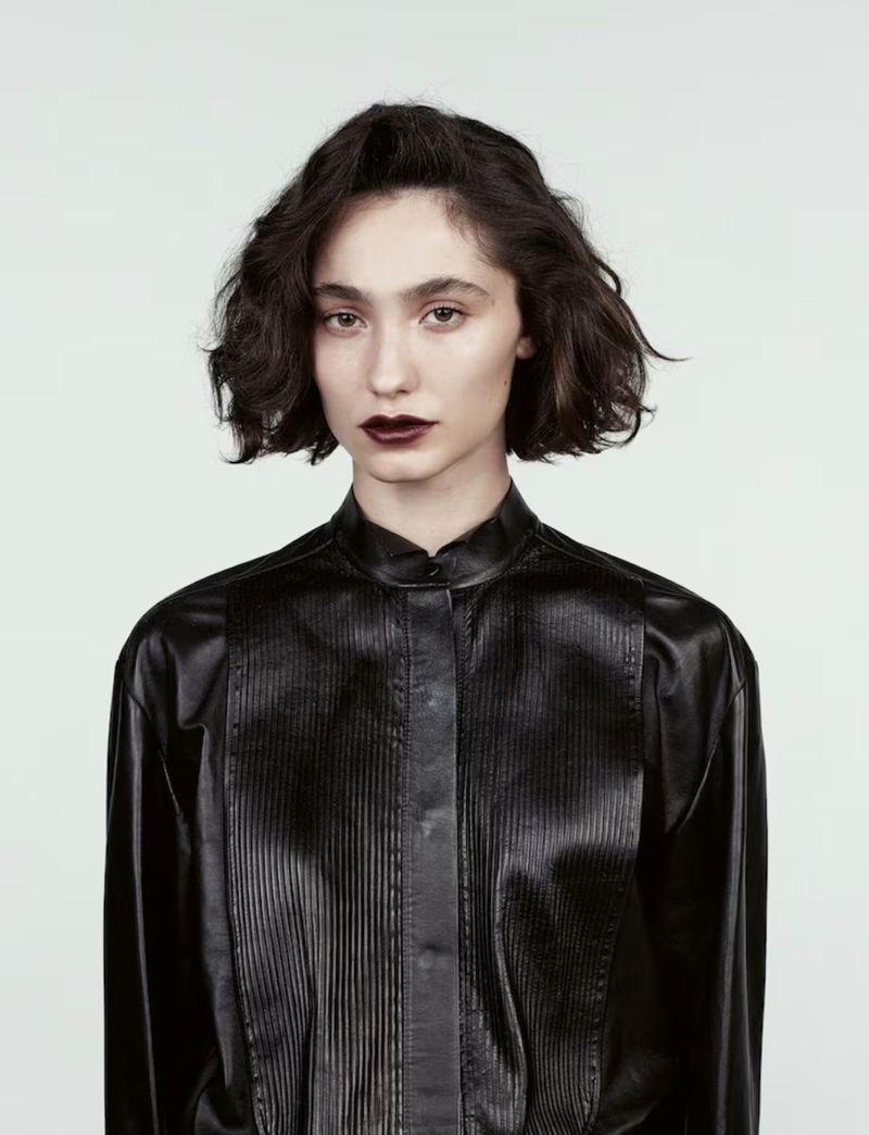 Portrait of a young woman with dark lipstick, wearing a black, long-sleeved, vertically-striped leather shirt against a white background.