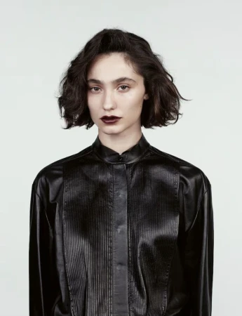 Portrait of a young woman with dark lipstick, wearing a black, long-sleeved, vertically-striped leather shirt against a white background.