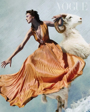 Fashion editorial of an adult female model in an orange dress posing with a white ram against a blue and white textured background.
