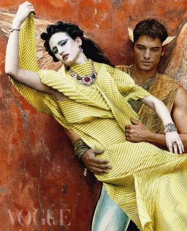 Editorial shot of a reclining woman in a yellow gown, embraced by a man in golden attire, posing against a textured ochre background.