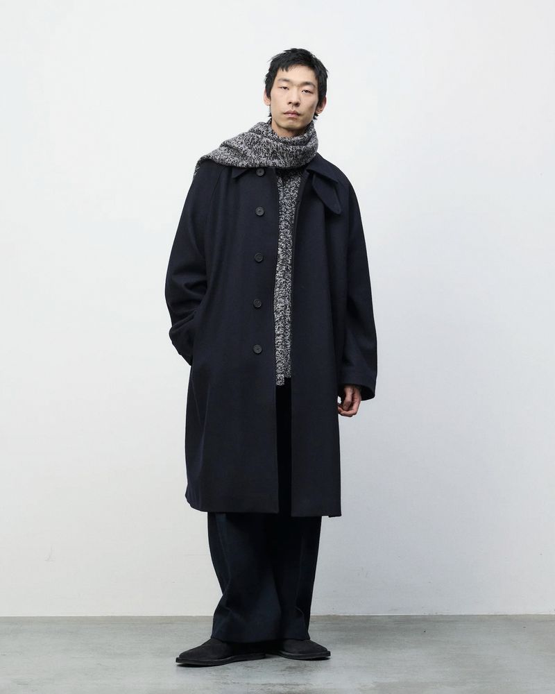 Adult Asian man in dark coat and knitted scarf stands against a plain background in a fashion style full shot.