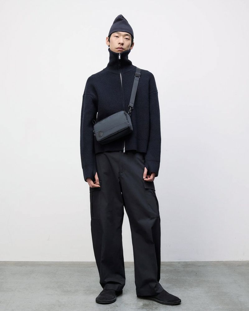 An Asian male model stands in a photo studio. He is wearing a navy beanie, knit sweater, black pants, and crossbody bag against a white backdrop.