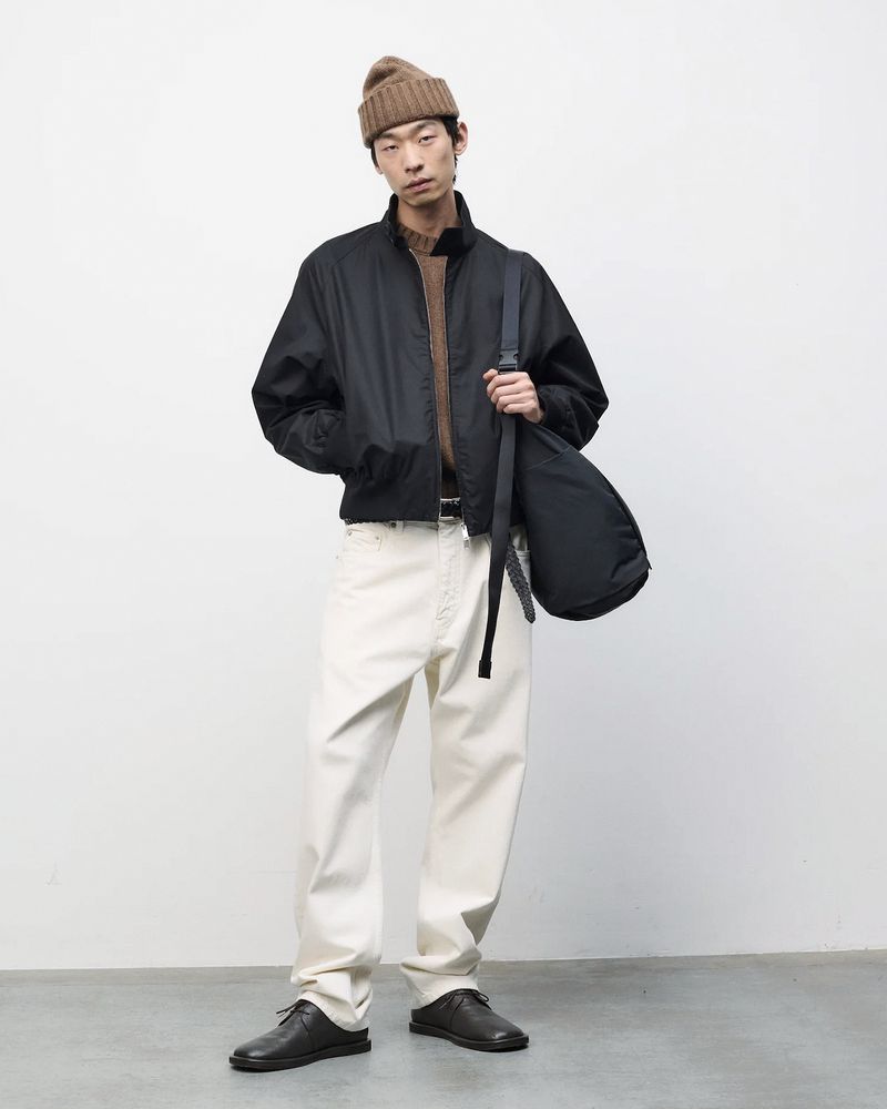 Full-length portrait of a young adult man wearing a beanie, jacket, light-colored jeans, and dark shoes against a plain background.