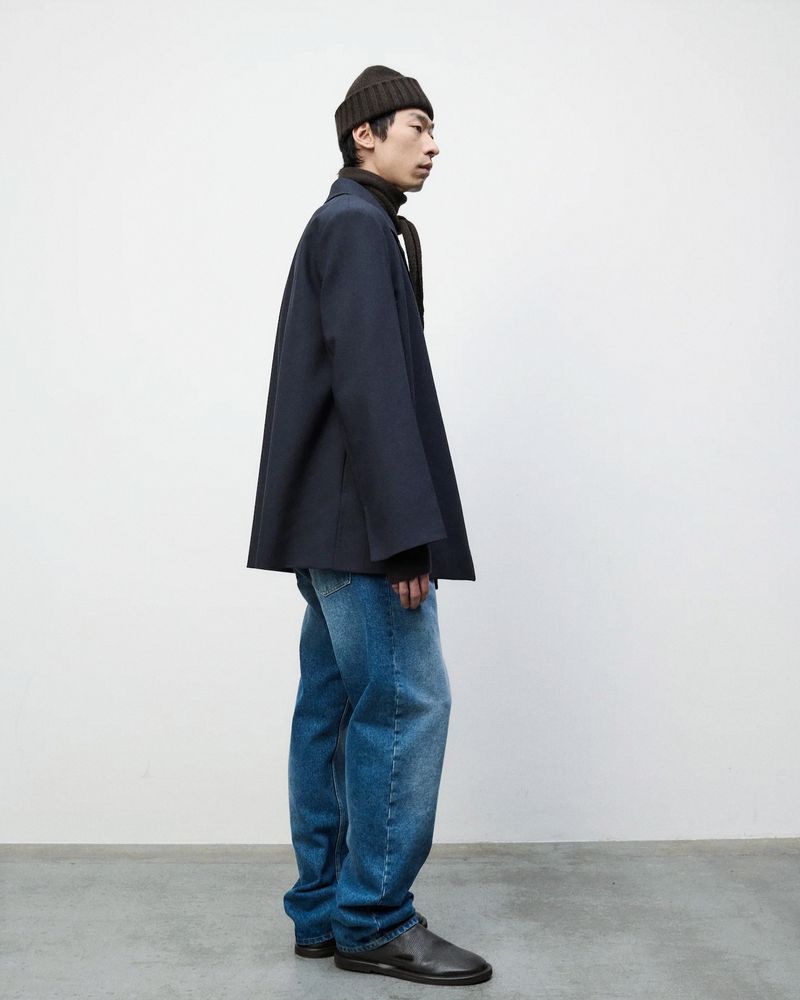 Side profile of an adult male wearing a navy blazer, jeans, a beanie, and leather shoes in a clean studio setting against a white backdrop.