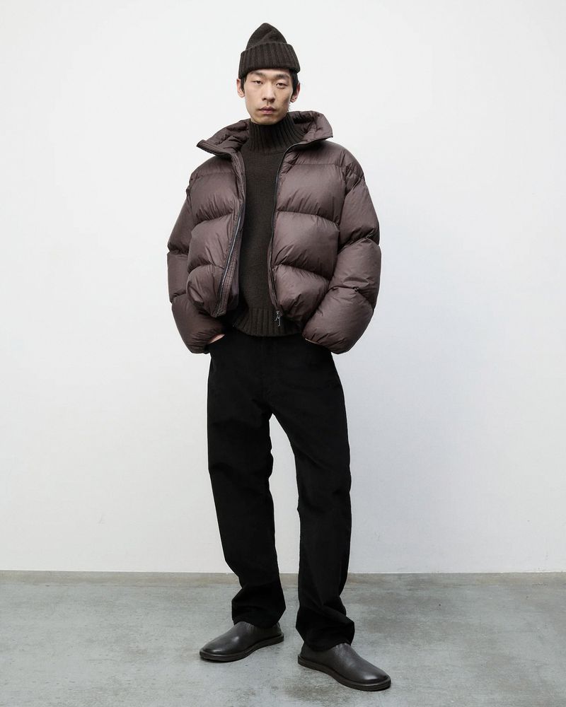 An adult male poses in a photo studio wearing a brown puffer jacket, dark brown turtleneck sweater, black pants, and black beanie.