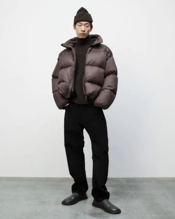 An adult male poses in a photo studio wearing a brown puffer jacket, dark brown turtleneck sweater, black pants, and black beanie.