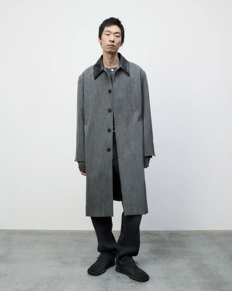 A slender adult male stands in front of a white wall, wearing a grey herringbone overcoat, grey shirt, black pants, and black shoes.