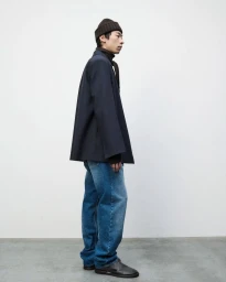 Model Zhuo Chen standing in profile wearing a dark navy structured jacket, blue jeans, and a dark beanie against a white studio wall.