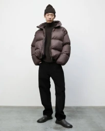 Model Zhuo Chen wearing a brown puffer jacket, dark turtleneck, black trousers, and beanie for Studio Nicholson Autumn Winter 25.
