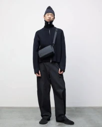 Model wearing a dark zip-up sweater, beanie, and trousers with a crossbody bag standing against a white seamless backdrop