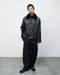 Model Zhuo Chen standing in a black textured leather jacket with a shearling collar and wide-leg trousers against a minimal wall.