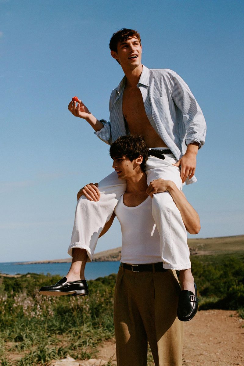 Two men in casual fashion pose in an outdoor coastal setting under a clear sky; one man sits on the shoulders of another, holding a fruit.