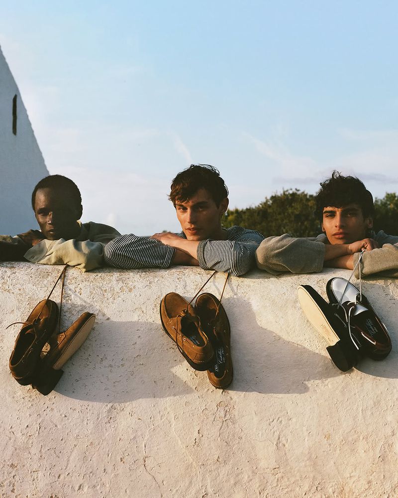 Three young men lean over a ledge with pairs of shoes dangling from the wall, bathed in soft, golden-hour sunlight against a clear sky.