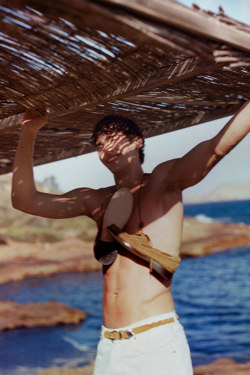Man with sandals on his chest stands near water wearing white shorts and a belt, holding a woven structure overhead on a sunny day.