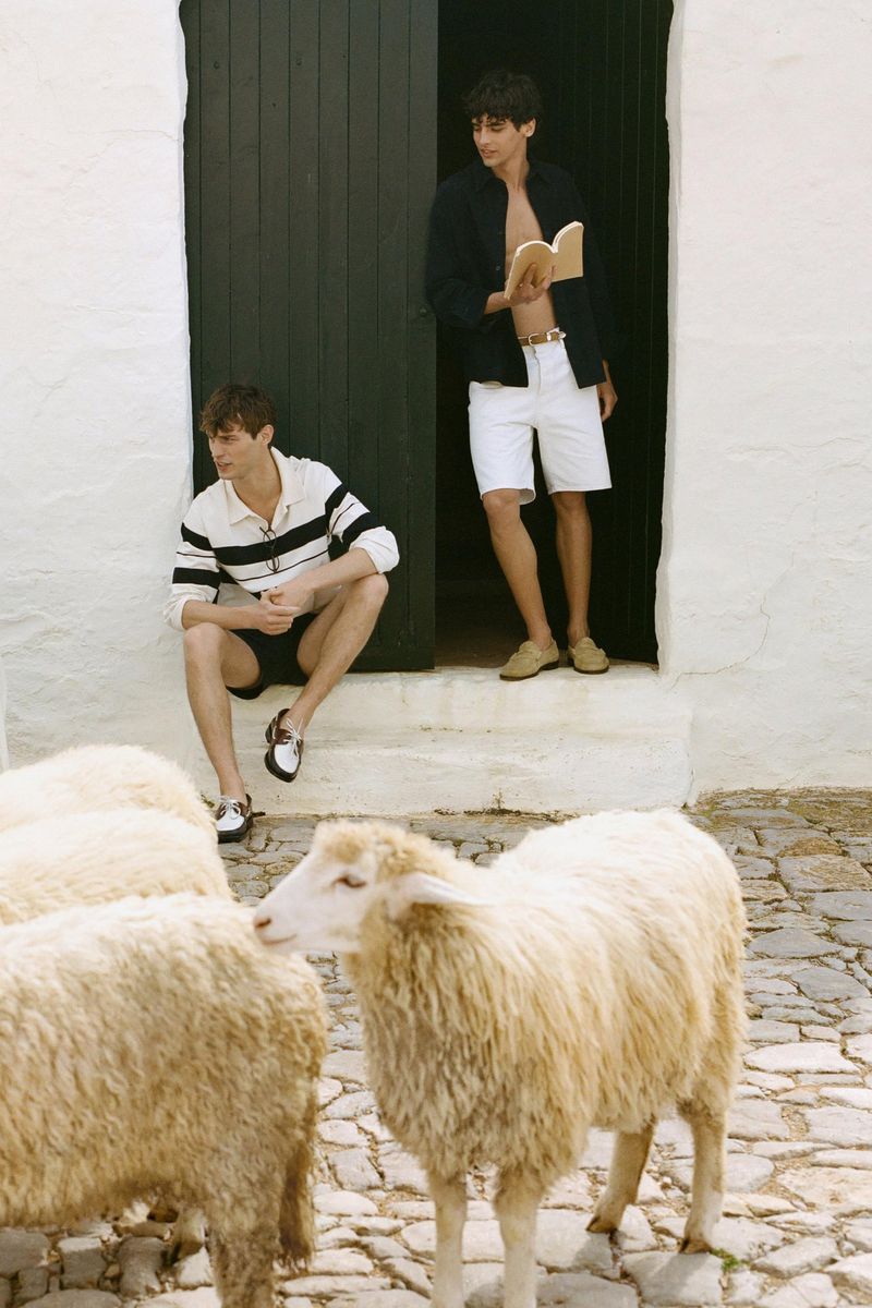 Two men pose outside white building with dark door near flock of sheep on a cobblestone path, wearing dark and light casual clothing.
