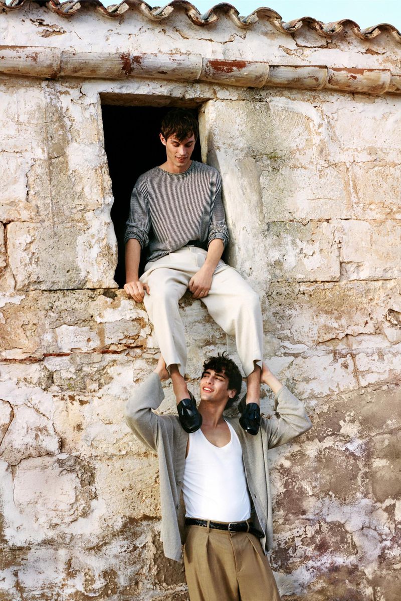 Two young adult males pose outdoors against a stone building with a window. One man sits on the window ledge while the other holds his feet.