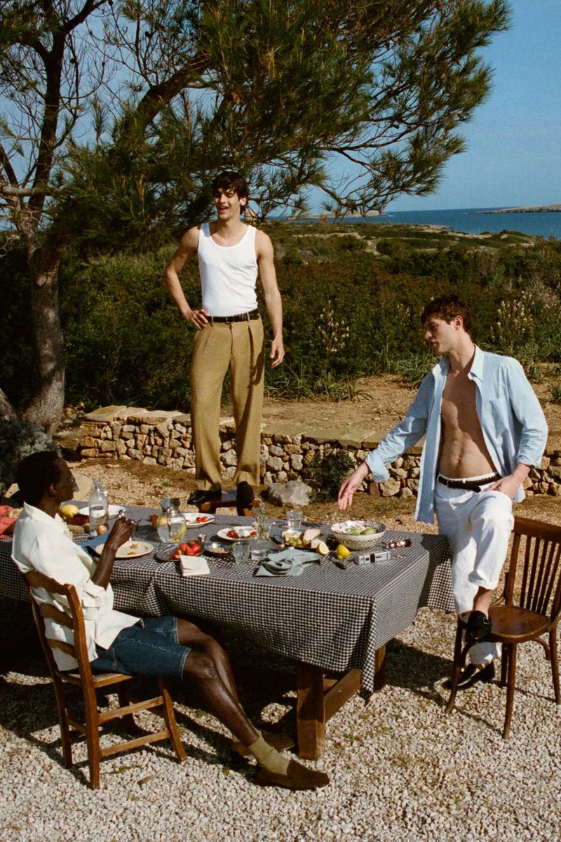 Three stylish men gather for an outdoor meal, sitting and standing by a table laden with food, under a tree on a sunny day.