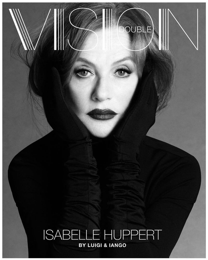 Black-and-white portrait of Isabelle Huppert in gloves, featuring dramatic dark lipstick, in front of a plain gray backdrop, editorial in style.