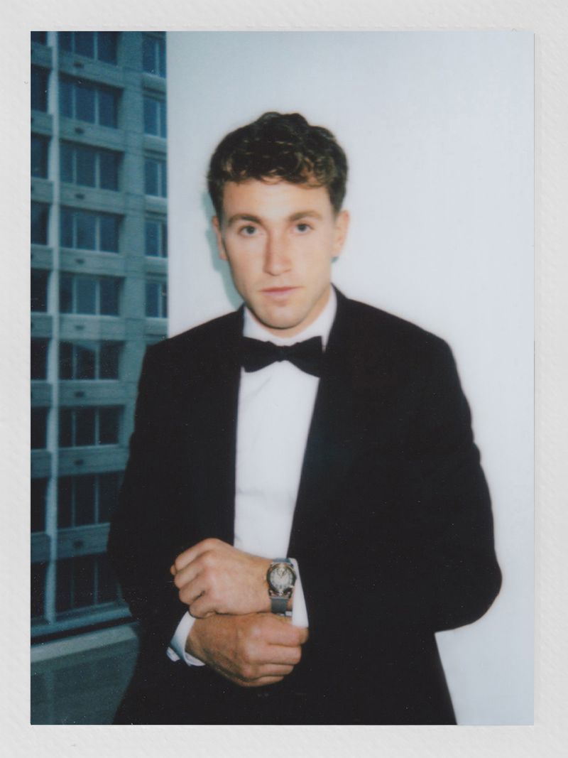 Portrait of a young adult male wearing a tuxedo and bow tie, posing indoors with a blurred city building in the background.
