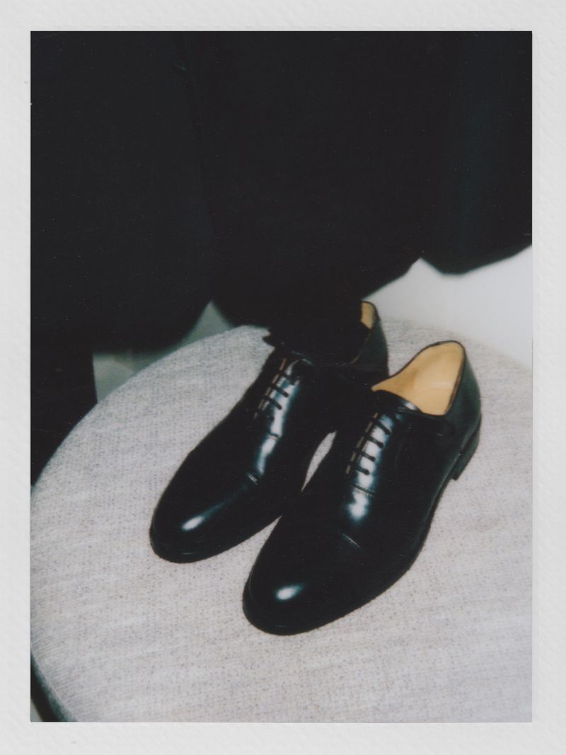 Close-up of a pair of glossy black leather dress shoes with laces sitting atop a gray cushioned stool with a black garment in background.