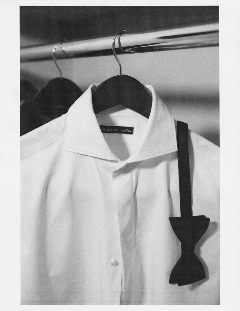 White dress shirt hangs on a wooden hanger with a black bow tie in a grayscale studio shot, showcasing fashion and menswear.