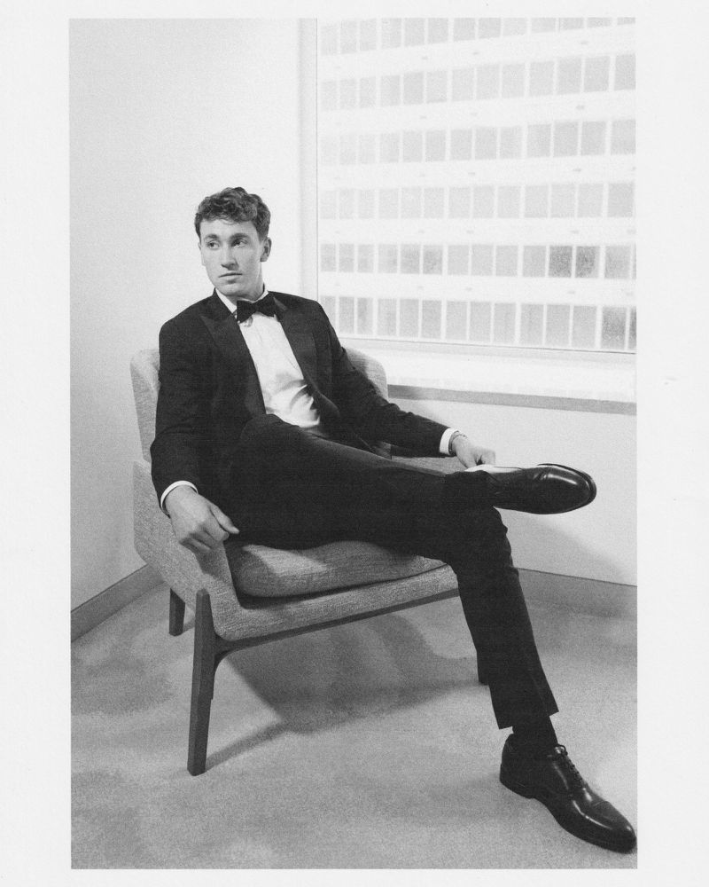 Black-and-white image of a man in a black tuxedo sitting in a chair with a large window and a white wall in the background.